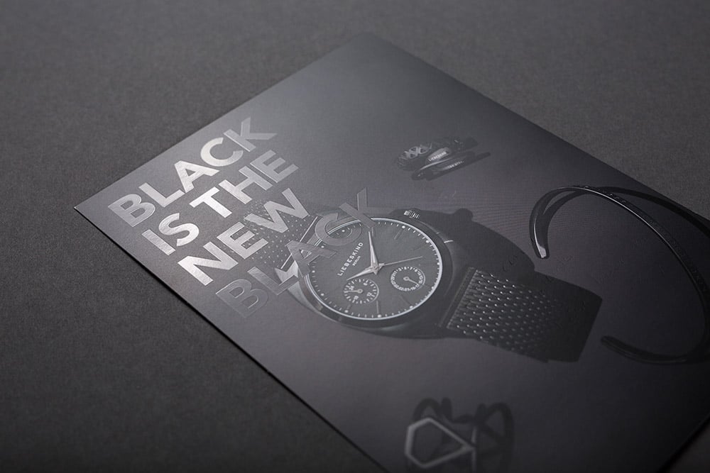Ein DIN A4 Flyer, der vollflächig schwarz bedruckt ist und mit einer partiellen Lackierung den Satz "black is the new black" veredelt hat