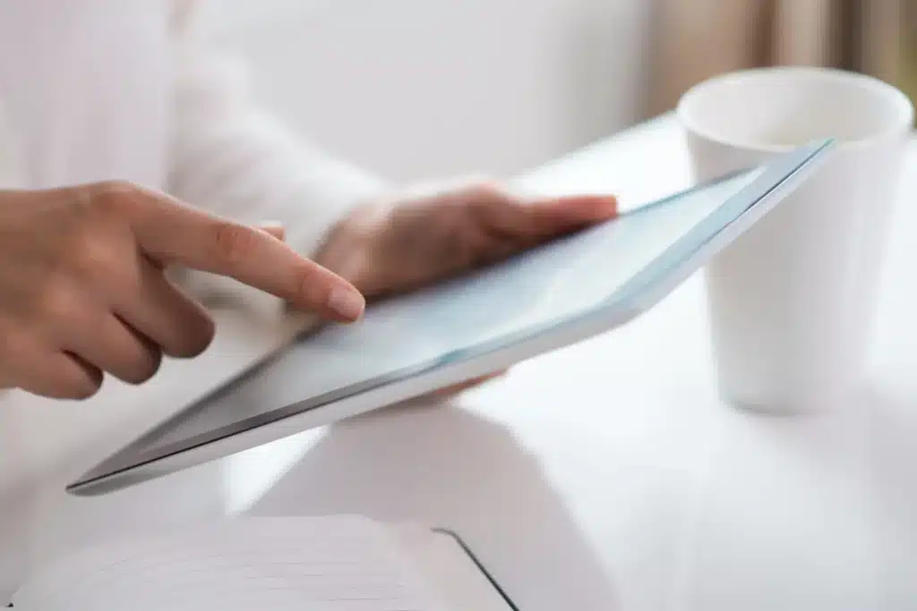 Detailaufnahme von zwei Händen, die ein Tablet halten. Eine Hand deutet mit dem Zeigefinger auf das Display. Die Umgebung ist schlicht in Weiß gehalten.